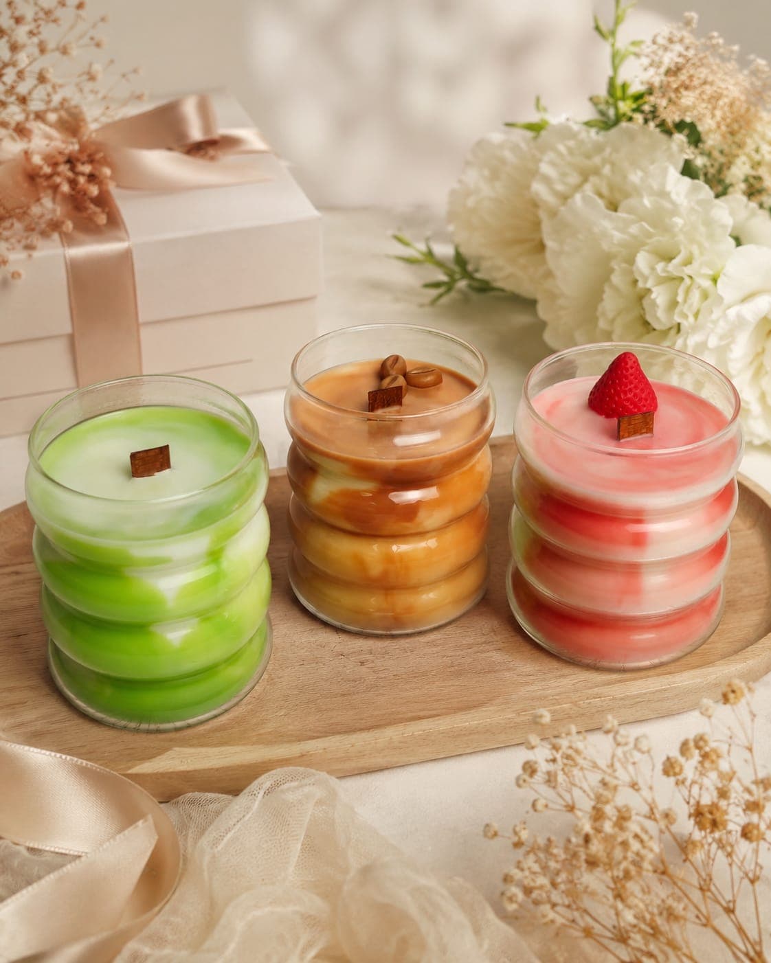 Las 3 velas de la Colección Bebidas en una bandeja de madera junto a una caja de regalo con lazo y un ramo de flores blancas