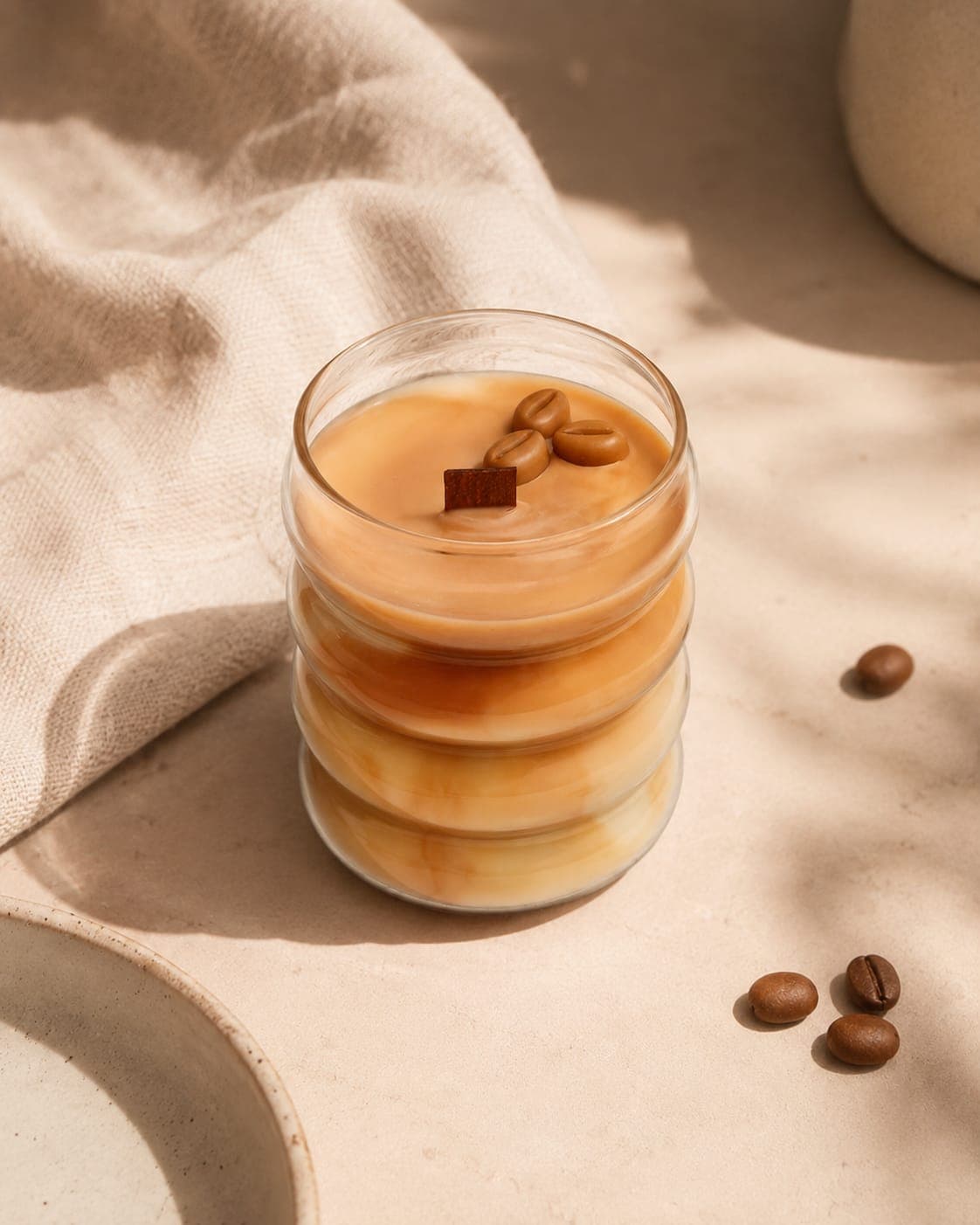 Vela Caramel Latte con luz natural sobre fondo beige, con un paño de lino y granos de café alrededor y las capas de cera visibles a través del vidrio ondulado
