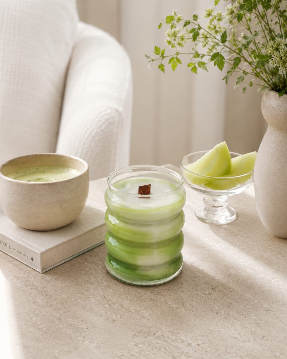 Vela Melon Matcha Latte sobre una mesa de piedra clara junto a una taza de matcha, un libro y una copa de cristal con trozos de melón